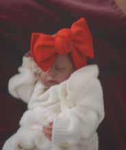 Newborn baby wrapped in white blanket with a large red bow on head against a dark background